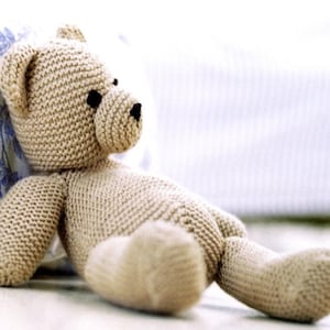 Op de afbeelding: Een handgemaakte, beige gebreide teddybeer met zwarte knoopogen en neus. De beer ligt languit. Het knuffel is gemaakt van een getextureerd garen, waardoor het een gezellige uitstraling heeft. Een klassiek, troostrijk kinderspeelgoed.