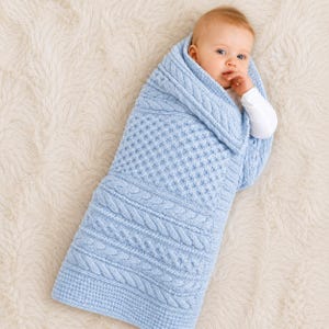 May include: A light blue knitted baby blanket featuring a textured cable knit pattern and a honeycomb stitch design. The blanket is wrapped around a baby wearing a white long-sleeved shirt, showcasing the intricate yarn work and soft blue color.