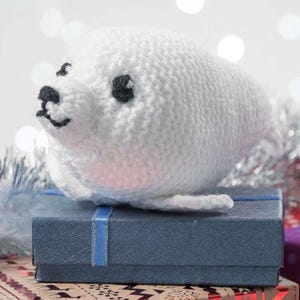 May include: A handmade white knitted seal toy with black eyes and a smiling mouth. The seal is resting on a blue gift box tied with a blue ribbon. The background features bokeh lights and silver tinsel.