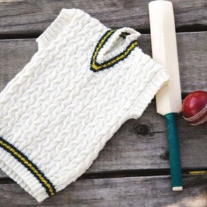 May include: Cream-colored cricket sweater with a cable knit design, accented with navy and yellow stripes. A wooden cricket bat with a green handle and a red cricket ball are next to the sweater, all on a weathered wooden surface.