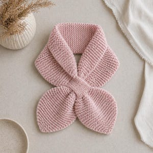 Puede incluir: Bufanda de punto rosa claro con un diseño único. La bufanda está hecha de un hilo suave y texturizado y presenta un diseño en bucle en la parte delantera. Se muestra sobre una superficie neutra con un jarrón y tela en el fondo.