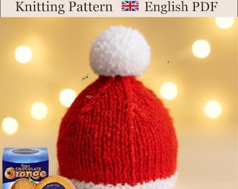 May include: A red knitted hat with a white pom-pom and a white brim. The text at the top reads: Chocolate Orange Cover Knitting Pattern English PDF. Next to the text is a British flag icon. A box of Terry's Chocolate Orange is visible.