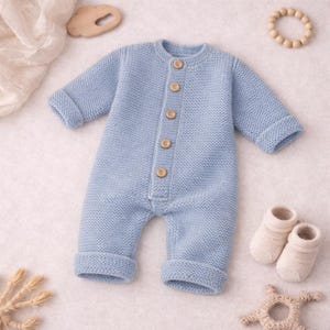 May include: A light blue knitted baby romper with long sleeves and wooden buttons down the front. The romper has folded cuffs on the sleeves and legs. A pair of cream-colored baby socks and wooden toys are also in the image.