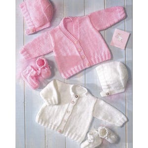 Può includere: Un set di vestiti per neonati fatti a mano in rosa tenue e bianco. Il set comprende un cardigan, un cappellino e delle scarpine. È presente anche un piccolo biglietto con un'illustrazione di cicogna. Gli articoli sono disposti su una superficie di legno azzurro chiaro.