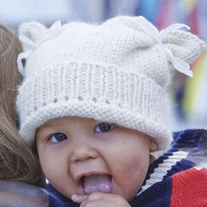 May include: A knitted white baby hat with two bow accents. The hat has a ribbed brim and is worn by a baby with dark eyes and their tongue sticking out. The baby is wearing a blue, red, and white striped shirt.