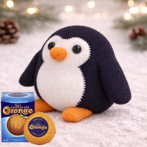 Puede incluir: Un pingüino tejido a mano con vientre blanco, cuerpo azul marino y pico y pies naranjas. Junto a él, una caja y una naranja de chocolate. La caja dice "Terry's Chocolate Orange" y la naranja de chocolate es visible.