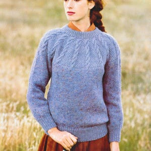Peut inclure: Un pull bleu tricoté avec un motif texturé sur l'empiècement et les manches. Le pull a une encolure ronde et des manches longues. Le modèle porte une jupe couleur rouille et une chemise marron à col en dessous.