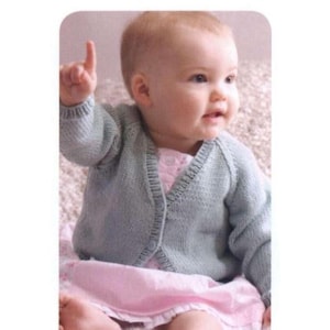 Op de afbeelding: Een baby draagt een lichtblauw gebreid vestje over een roze jurk. De baby zit en wijst met één vinger omhoog. Het vestje heeft een V-hals en een knoopsluiting. De outfit is zacht en comfortabel.