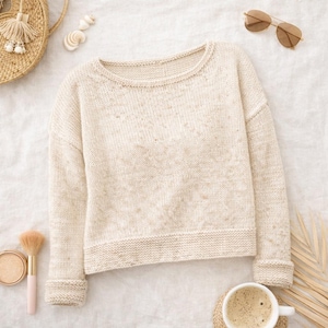 May include: A cream-coloured knitted sweater with a round neckline and long sleeves. The sweater features a speckled pattern and ribbed cuffs and hem. The image also includes a straw bag, sunglasses, makeup, and a cup of coffee, suggesting a relaxed, casual theme.