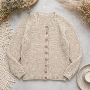 May include: A beige button-down cardigan with a textured knit pattern. The sweater features a round neckline, long sleeves, and wooden buttons. The garment is displayed on a neutral surface with decorative elements.