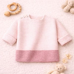 May include: A pale pink baby sweater with a darker pink hem and three buttons on the shoulder. The sweater has long sleeves with folded cuffs. A wooden teething ring and two teddy bears are in the background.