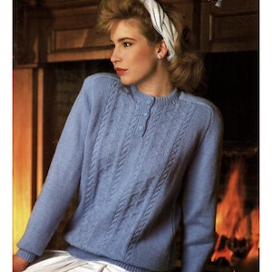 May include: A light blue cable-knit sweater with a crew neck and three small buttons. The long-sleeved sweater has a textured pattern and ribbed cuffs. A white headband is worn on the head.