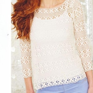 May include: Cream-colored crochet top with long sleeves. The top features a scoop neckline and intricate openwork patterns throughout the design. The sleeves are three-quarter length, and the bottom hem has a scalloped edge. The top is paired with light blue jeans.