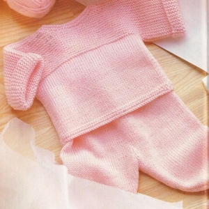 Może przedstawiać: Jasnoróżowy, dzianinowy zestaw dla dziecka, składający się ze sweterka i spodni, jest prezentowany na drewnianej powierzchni. Sweter ma długie rękawy i okrągły dekolt. W tle widoczna jest kulka różowej włóczki. Zestaw wykonany jest z miękkiej dzianiny.