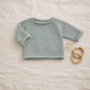 Puede incluir: Un suéter de bebé de punto azul claro con mangas largas y cuello redondo. El suéter está hecho de un hilo suave y texturizado. Un mordedor de madera está al lado del suéter.