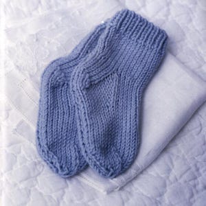 May include: A pair of light blue knitted socks, resting on a white, textured surface. The socks are made of a thick yarn, with visible knit stitches. The socks are folded neatly, suggesting they are new or unused.