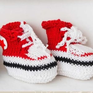 Op de afbeelding: Een paar rood, wit en zwart gebreide babyschoentjes. De schoentjes zijn ontworpen om op hoge sneakers te lijken, compleet met witte veters en een sterembleem aan de zijkant. De kleurstelling is rood, wit en zwart.