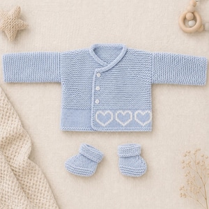 Peut inclure: Ensemble de vêtements pour bébé en tricot bleu clair, comprenant un cardigan avec des cœurs blancs et des chaussons assortis. Le cardigan a une fermeture à boutons et un col. L'ensemble est en fil doux et texturé.