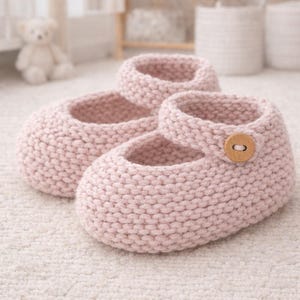 Puede incluir: Un par de zapatos de bebé de punto rosa claro, con una correa Mary Jane y un cierre de botón de madera. Los zapatos están hechos de un hilo suave y texturizado y están diseñados para bebés. Los zapatos se exhiben sobre una superficie blanca.