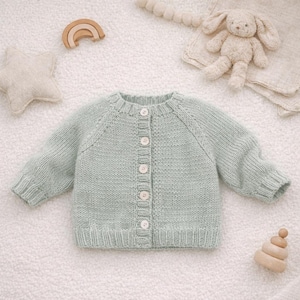 Könnte beinhalten: Hellgrüne Baby-Strickjacke mit langen Ärmeln und Knopfleiste. Der weiche, strukturierte Pullover ist von Babyspielzeug umgeben, darunter ein Stoffhase, ein Stern und ein Holzregenbogen.