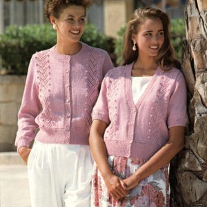 Puede incluir: Dos mujeres con cárdigans de punto rosa a juego. Uno tiene mangas largas, el otro cortas. Ambos tienen botones en la parte delantera y patrones de punto decorativos. Una lleva pantalones blancos, la otra una falda floral. Los cárdigans son de color rosa suave.