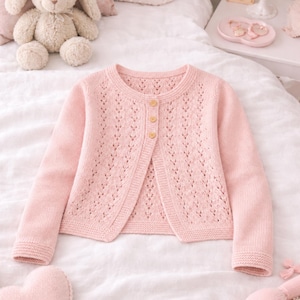 May include: A pale pink cardigan with an openwork pattern. The long-sleeved, knitted sweater has a round neck and is fastened with three wooden buttons. The garment is displayed on a white surface.
