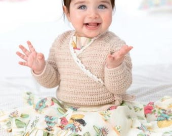 Puede incluir: Un bebé sonriente con un cárdigan envolvente de crochet beige claro con ribete blanco. El bebé también lleva una falda con estampado floral y dobladillo con volantes. La falda presenta ilustraciones de personas y plantas.