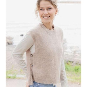 Puede incluir: Un chaleco de punto beige con cuello redondo y botones laterales. El chaleco se lleva sobre una camisa de manga larga de color claro. La modelo sonríe y está de pie al aire libre cerca de una masa de agua.