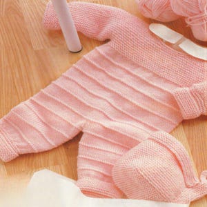 May include: A light pink knitted baby outfit, including a one-piece suit and a matching cap. The suit features ribbed detailing across the chest and legs. The outfit is made of soft yarn, perfect for a newborn.