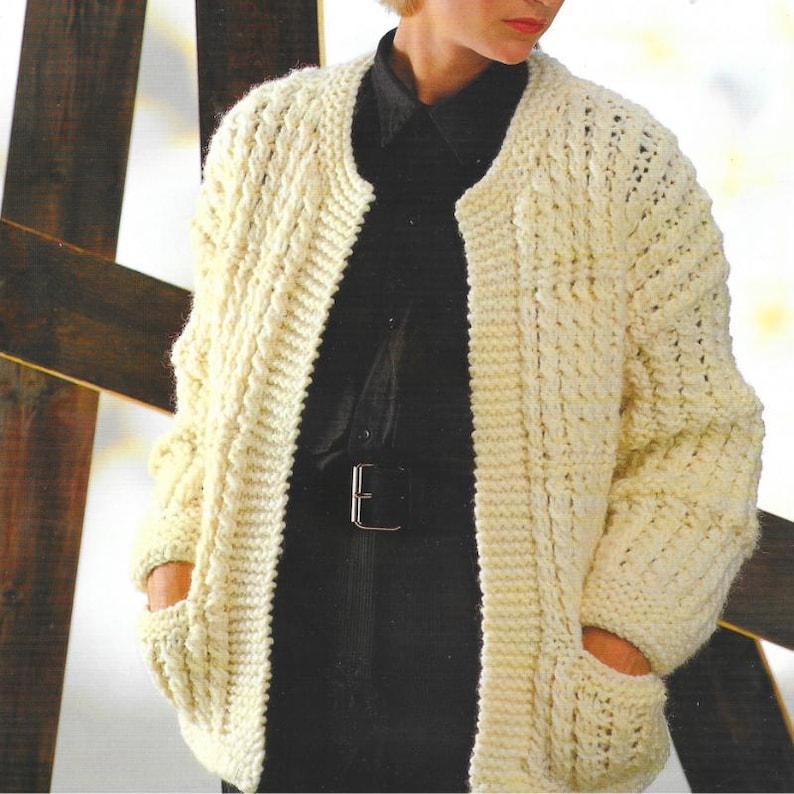 May include: Cream-colored, open-front cardigan with a textured knit pattern. The sweater has long sleeves and two front pockets. The person in the image is wearing a black shirt and belt underneath the cardigan.