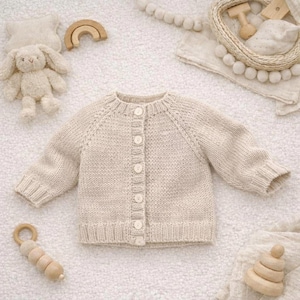 Puede incluir: Un cárdigan de bebé color crema con botones de punto. El suéter de manga larga tiene un cuello redondo y está rodeado de juguetes para bebés, incluyendo un conejito de peluche, un arcoíris de madera y una sonaja de madera.