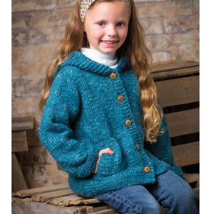 Könnte beinhalten: Eine türkisfarbene Kapuzenstrickjacke mit Holzknöpfen und Taschen. Die Jacke wird von einer Person mit langen blonden Haaren und Jeans getragen. Die Jacke ist aus einem strukturierten Garn gefertigt und hat ein gemütliches, handgemachtes Aussehen.