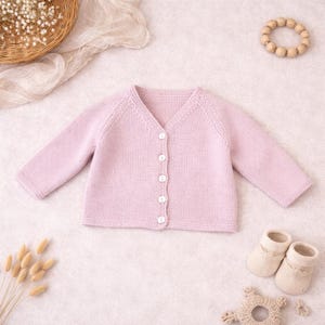 May include: A light pink, button-down baby cardigan with long sleeves. The knitted sweater has a V-neck and white buttons. A pair of cream-colored baby booties and a wooden teething ring are also visible.