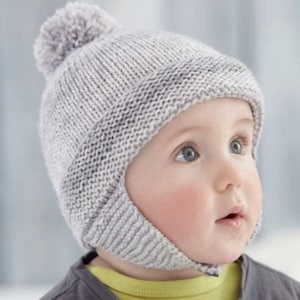 Puede incluir: Un gorro de bebé de punto gris claro con orejeras y un pompón en la parte superior. El gorro está hecho de un hilo suave y texturizado y está diseñado para mantener la cabeza y las orejas del bebé calientes. El bebé lleva una camiseta gris y otra amarilla debajo.
