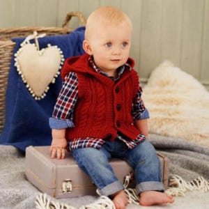 May include: A baby wearing a red hooded cable-knit vest over a plaid shirt and blue jeans. The baby is sitting on a small, vintage suitcase. The outfit is a classic, cozy style for infants.
