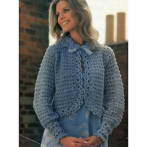 Peut inclure: Une veste en crochet bleu clair avec un col et des manches longues. La veste présente un motif ajouré délicat et une fermeture à nœud au niveau de l'encolure. La veste est portée sur une robe bleue.