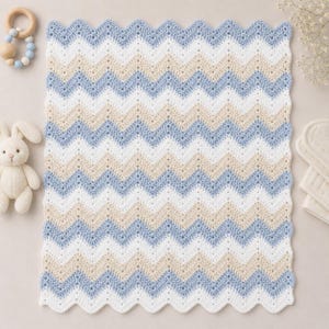 May include: A crocheted baby blanket featuring a chevron pattern in white, pale blue, and beige. The blanket has a scalloped edge. A wooden baby rattle and a stuffed bunny are next to the blanket.