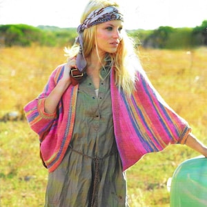 May include: A pink, blue, and orange striped knit cardigan with wide sleeves. The cardigan is worn over a green dress with a brown belt. A patterned scarf is worn as a headband. The background is a field of dry grass.