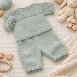 May include: A light green knitted baby outfit, including a long-sleeved top and pants. The outfit is made of soft yarn and has a ribbed cuff design. The outfit is displayed with baby toys, including wooden blocks and a stuffed bunny.