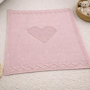 Può includere: Una coperta per neonati rosa chiaro lavorata a maglia con un motivo a cuore al centro. La coperta ha un bordo decorativo con un bordo smerlato. Un piccolo orsacchiotto è in un cestino di vimini sullo sfondo.