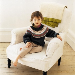 May include: A child in a dark grey sweater with a patterned yoke, seated in a white armchair. The sweater features a colourful geometric design around the neck and shoulders. The child wears beige trousers. A cream blanket with a green trim is draped over the chair.