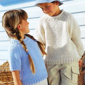 Può includere: Due bambini con maglioni lavorati a mano. Uno indossa un maglione azzurro a maniche corte, l'altro un maglione bianco sporco a maniche lunghe. Si sorridono. Lo sfondo è un muro bianco.