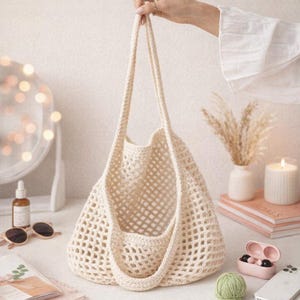 May include: Cream-coloured, open-weave crochet tote bag with long handles. The bag is held up by a hand. The bag is sitting on a white surface with other items, including sunglasses, a candle, and a book.