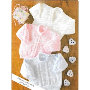 May include: Three knitted baby sweaters in white, pink, and light blue, with a delicate lace pattern. The sweaters are arranged on a wooden surface with heart-shaped buttons and a small package of floral-printed fabric. The text "PREMATURE SIZE INCLUDED" is visible.