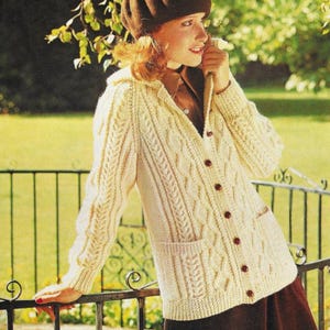 May include: Cream-colored, hooded cardigan sweater with a cable knit design. The sweater has a button-up front, two front pockets, and ribbed cuffs. The model is wearing a brown beret and a brown shirt.