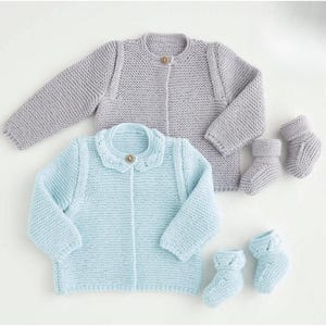 Puede incluir: Dos cárdigans de bebé de punto, uno azul claro y otro gris, con patucos a juego. Los cárdigans tienen cierre de botón y manga larga. El cárdigan azul claro tiene un cuello con volantes. Los patucos están doblados en la parte superior.