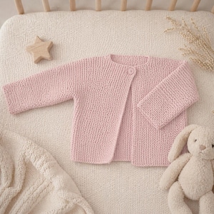 Puede incluir: Cárdigan de bebé tejido de color rosa claro con un solo botón. El cárdigan se muestra sobre una superficie texturizada de color crema, junto con una estrella de madera y un conejito de peluche.