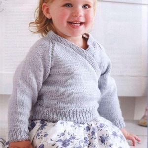 May include: A pale blue knitted wrap-style cardigan with long sleeves. The cardigan features a V-neck and a ribbed hem. The child is wearing a floral print skirt. The cardigan is made of a soft, comfortable material.