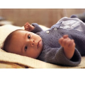 May include: A baby in a blue knitted sweater with two white buttons, resting on a cream blanket. The baby is looking at the camera with its arms raised.