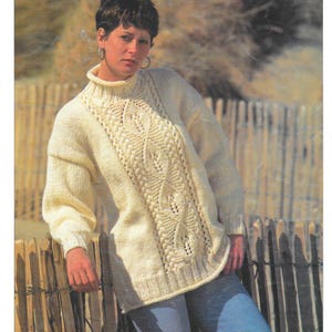 Może przedstawiać: Kremowy sweter z dzianiny z wysokim dekoltem i misternymi detalami warkoczowymi z przodu. Sweter ma długie rękawy i luźny krój. Osoba ma na sobie niebieskie dżinsy i opiera się o drewniany płot.